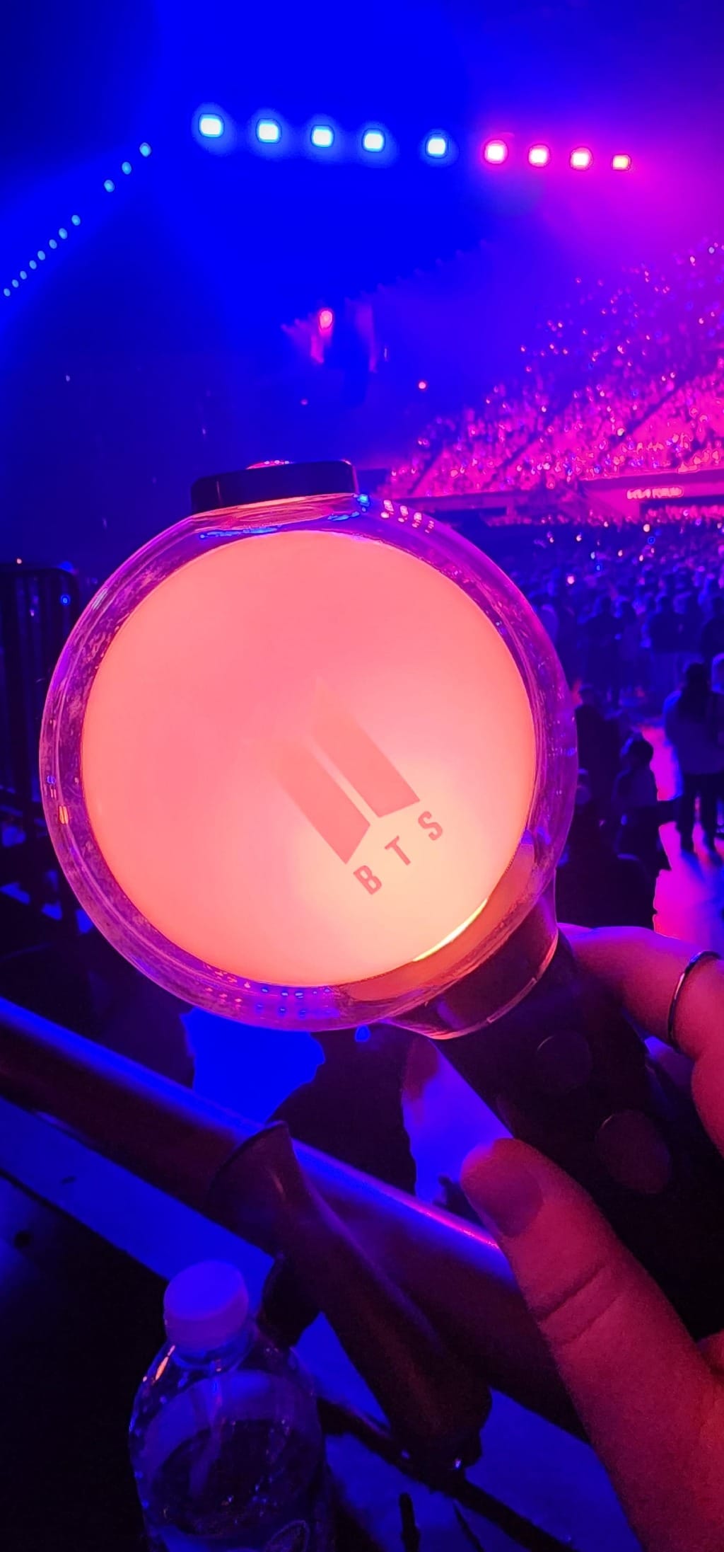 A BTS ARMY lightstick, lit up light orange, being held in front of a crowd in a stadium lit with red and blue.