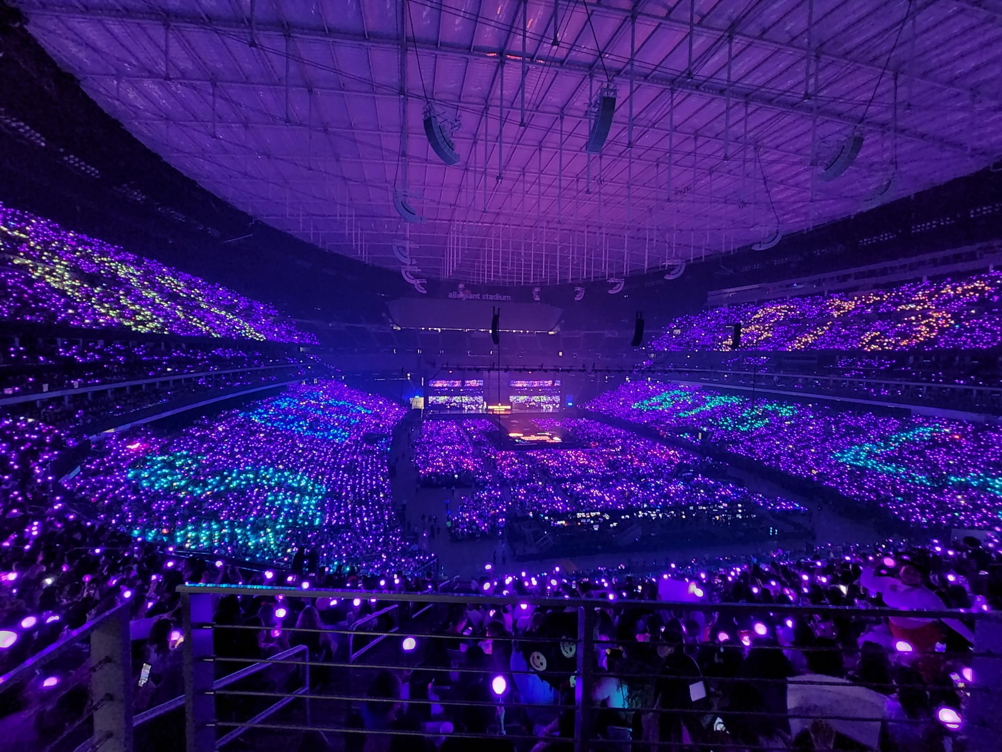 A stadium with purple lighting from stands as everyone in audience holds lightsticks, stage far across other side of stadium.