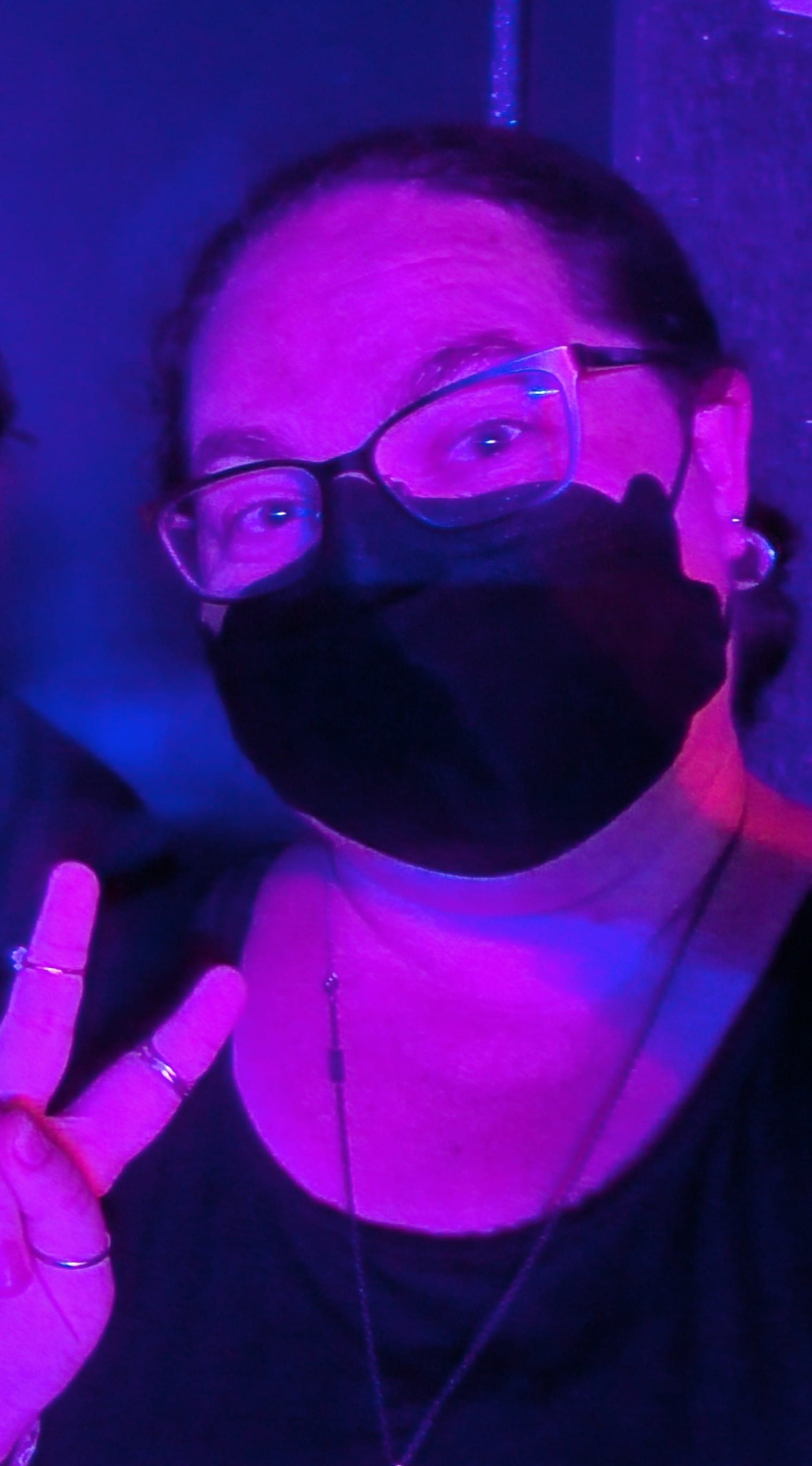 White woman wearing a black face mask and glasses making a peace sign. Everything is tinted rosy pink from the lighting.