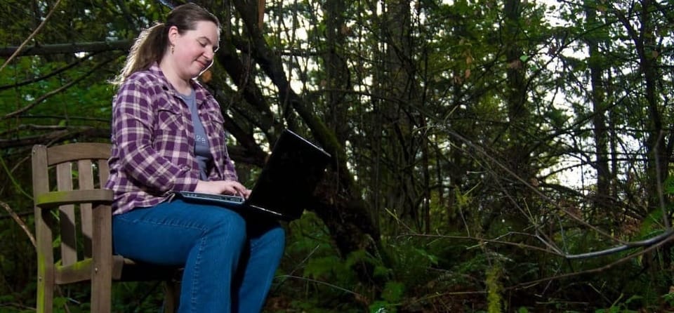 White woman with brown hair sitting on a wooden chair in a mossy woods with a laptop on her lap.