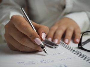 Two hands on a notebook, one holding a pen writing in the notebook