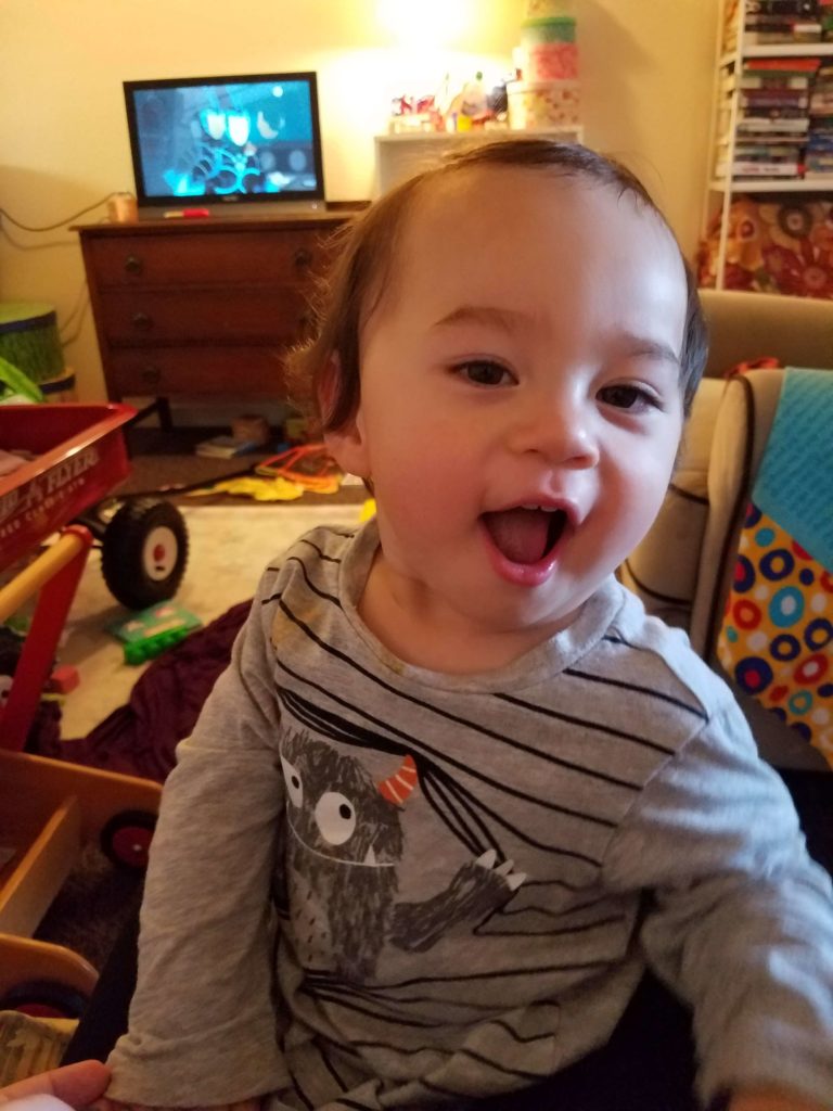A toddler smiling open mouthed at the camera with a monster shirt on.