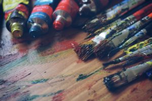 Paint tubes and paint brushes on a wooden table covered in different colored paints.