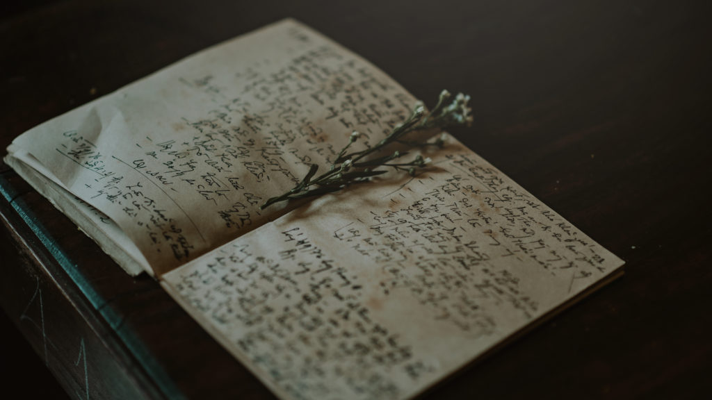 An open notebook on a dark wood desk with cursive writing in dark pen and a flower in the center of the book.